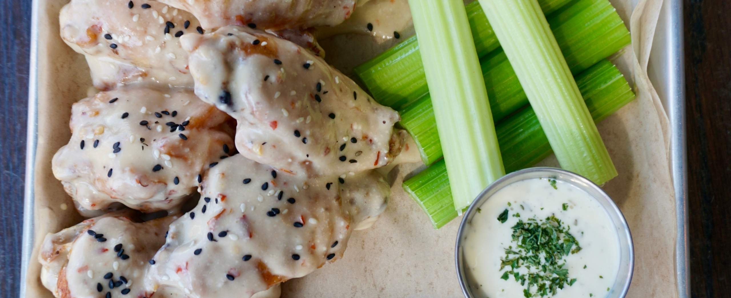 Chicken wings coated in a creamy white sauce with black sesame seeds, served with celery sticks and a side of ranch dressing topped with chopped herbs on a tray.