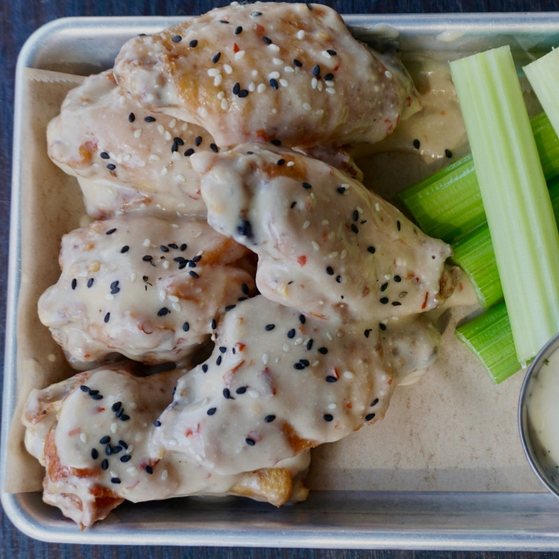 Chicken wings covered in a creamy sesame sauce, topped with black and white sesame seeds, served on a metal tray with celery sticks and a cup of dipping sauce on the side.