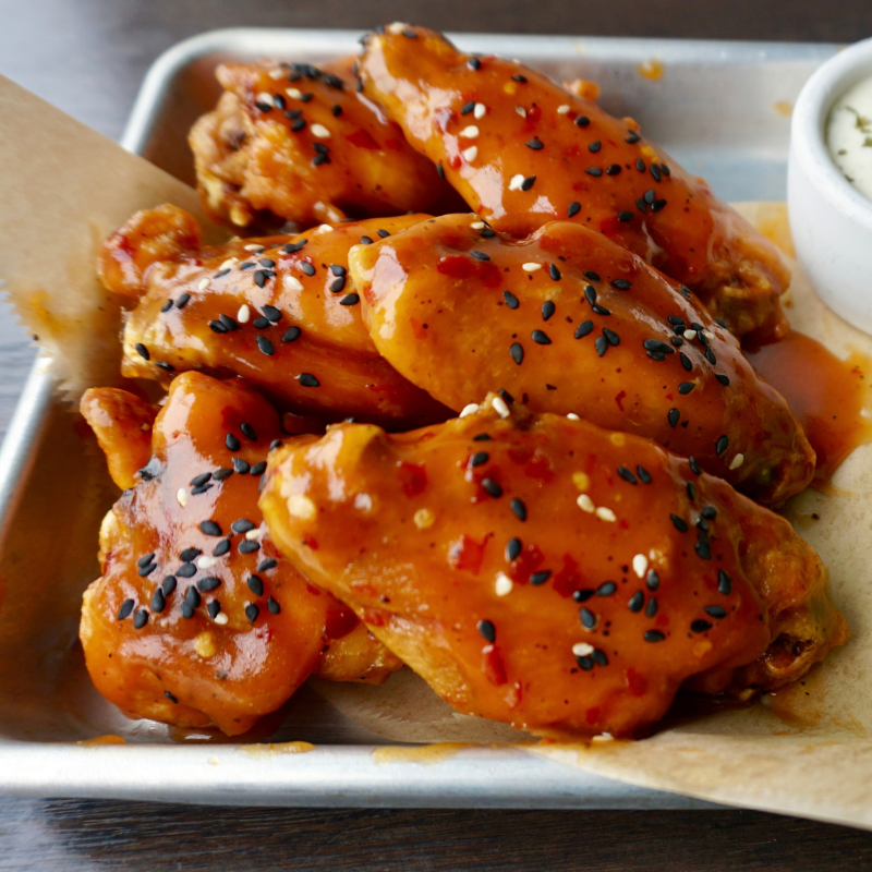 A tray of glazed chicken wings topped with black and white sesame seeds, served on parchment paper with a side of dipping sauce in a small white cup.