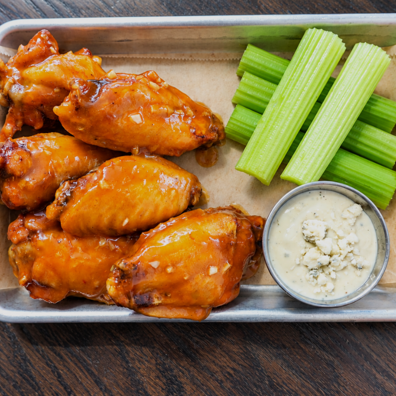 A metal tray with six saucy chicken wings, celery sticks, and a small cup of blue cheese dressing on parchment paper, on a dark wooden table.
