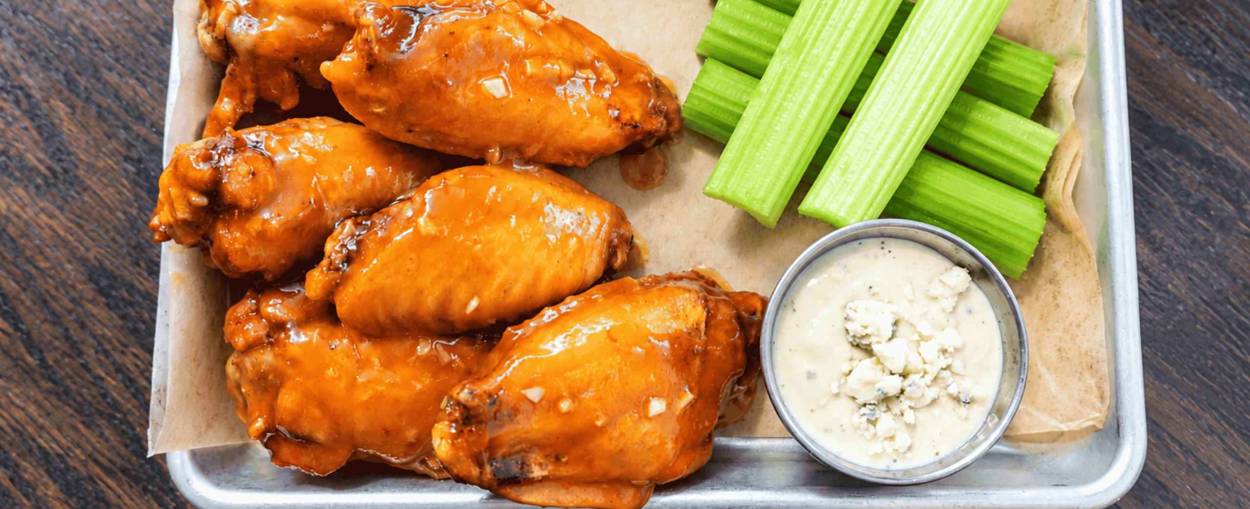 A tray with buffalo chicken wings, celery sticks, and a small cup of blue cheese dressing on a wooden table.