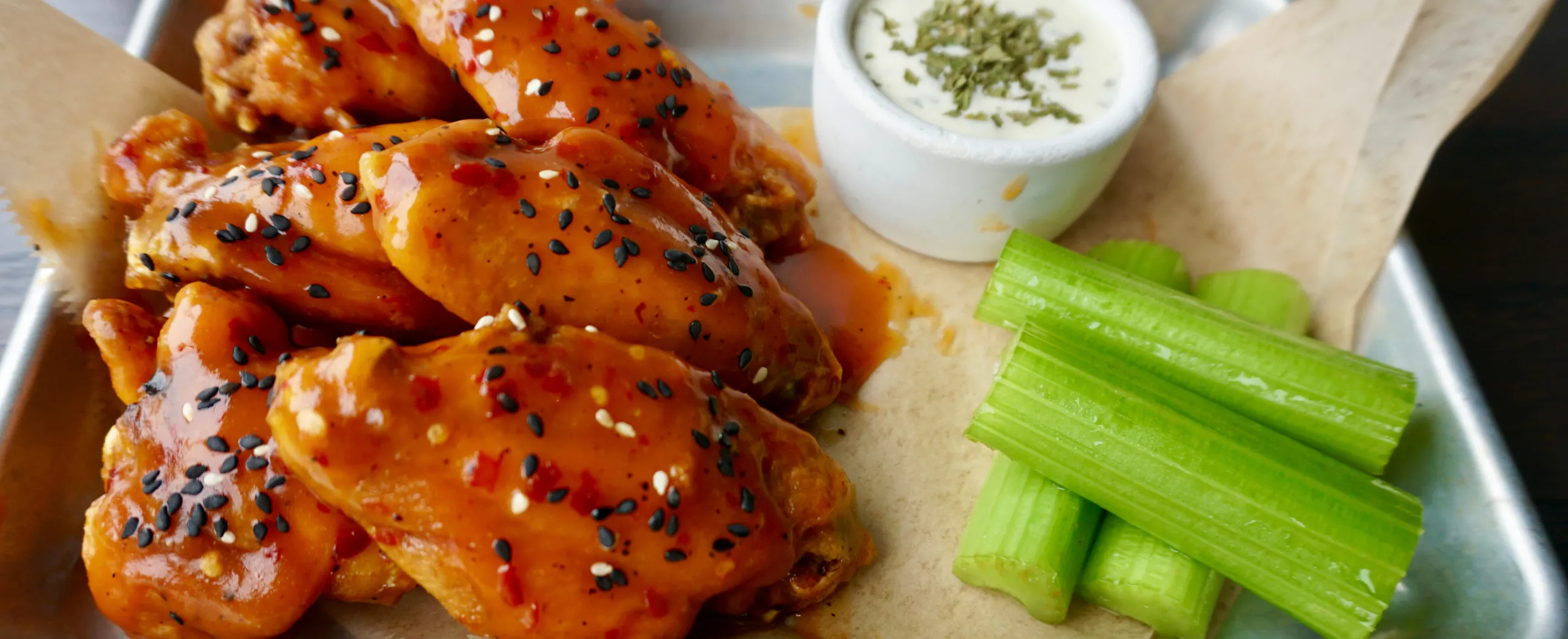 Buffalo chicken wings topped with sesame seeds and red pepper flakes, served with celery sticks and a cup of ranch dipping sauce on a metal tray lined with parchment paper.