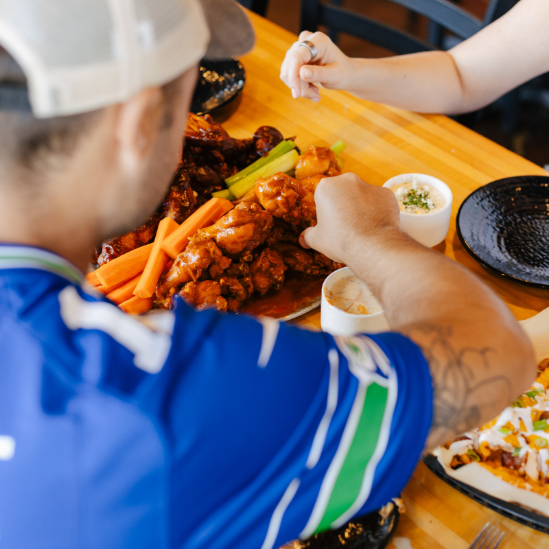 Two people sit at a table with chicken wings, celery, carrot sticks, and dipping sauces. One person wears a sports jersey and white cap; their hand reaches for food. The other person holds a carrot stick.